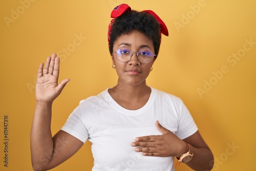 Young african american woman standing over yellow background swearing with hand on chest and open palm, making a loyalty promise oath