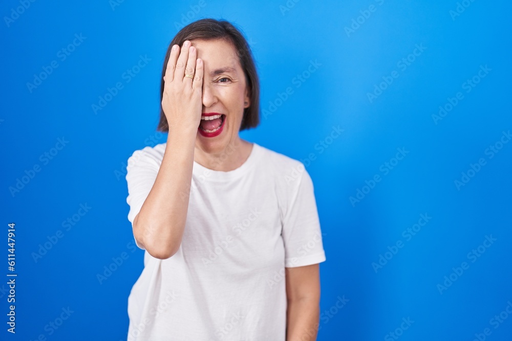 Middle age hispanic woman standing over blue background covering one eye with hand, confident smile on face and surprise emotion.