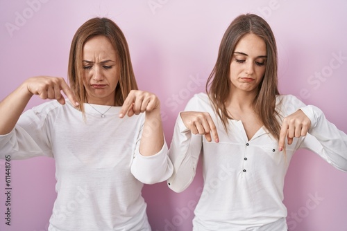 Middle age mother and young daughter standing over pink background pointing down looking sad and upset  indicating direction with fingers  unhappy and depressed.