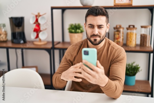 Young hispanic man using smartphone sitting on table at home