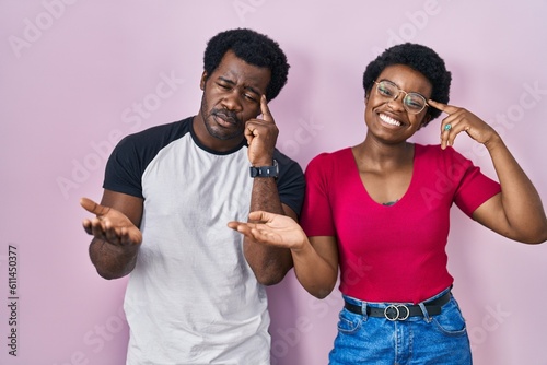 Young african american couple standing over pink background confused and annoyed with open palm showing copy space and pointing finger to forehead. think about it.