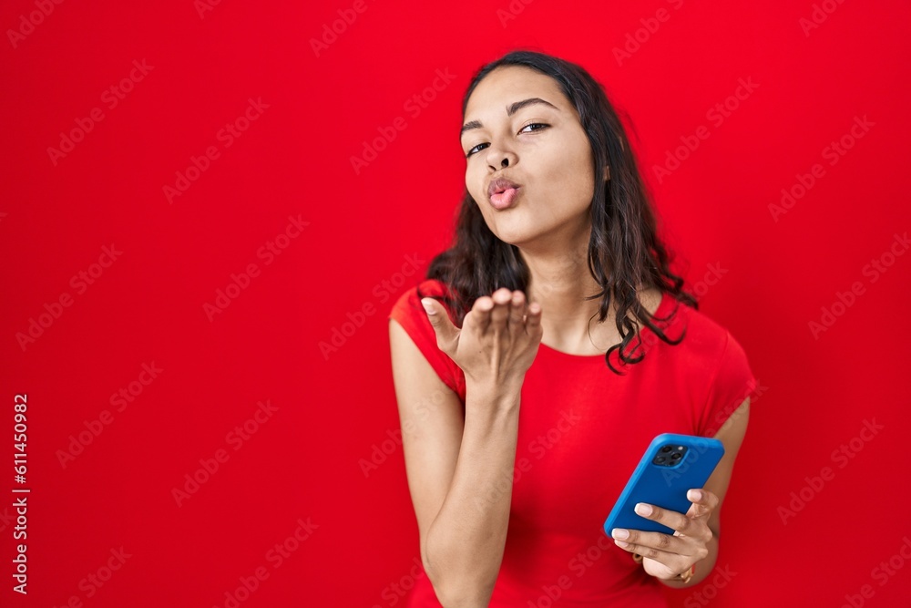 Young brazilian woman using smartphone over red background looking at the camera blowing a kiss with hand on air being lovely and sexy. love expression. Young brazilian woman using smartphone over red background looking at the camera blowing a kiss with hand on air being lovely and sexy. love expression.