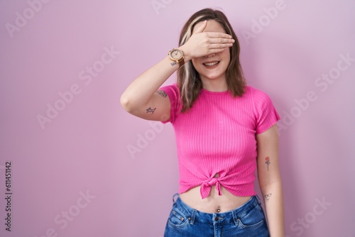 Blonde caucasian woman standing over pink background smiling and laughing with hand on face covering eyes for surprise. blind concept.