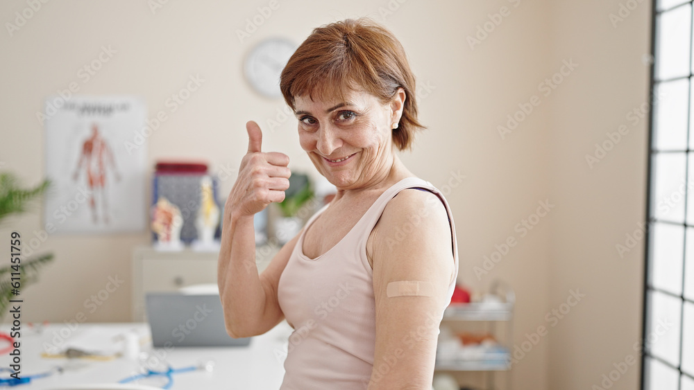 Mature hispanic woman doing thumbs up with band aid on arm at clinic Mature hispanic woman doing thumbs up with band aid on arm at clinic