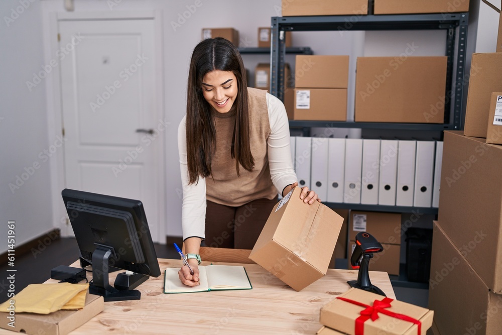 Young beautiful hispanic woman ecommerce business worker writing on notebook holding package at office Young beautiful hispanic woman ecommerce business worker writing on notebook holding package at office