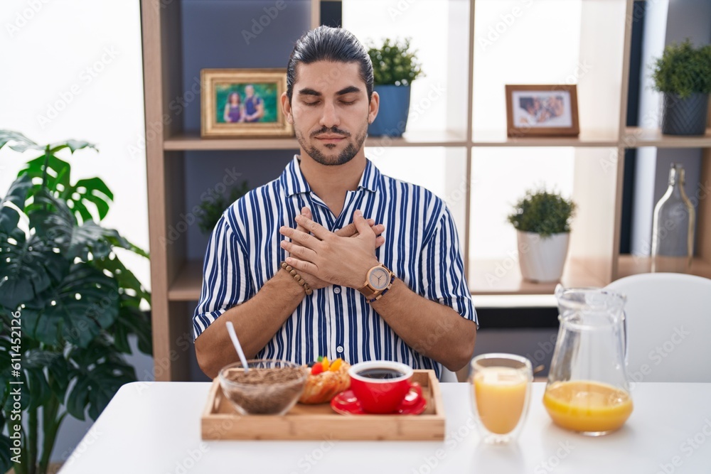 Hispanic man with long hair sitting on the table having breakfast smiling with hands on chest with closed eyes and grateful gesture on face. health concept. Hispanic man with long hair sitting on the table having breakfast smiling with hands on chest with closed eyes and grateful gesture on face. health concept.
