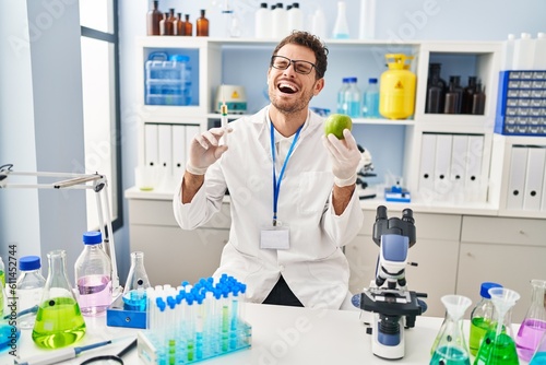 Young hispanic man working at scientist laboratory holding apple smiling and laughing hard out loud because funny crazy joke.
