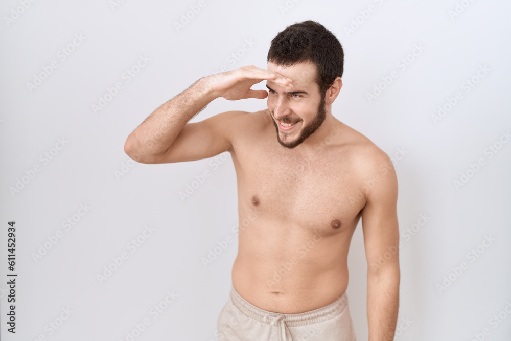 Young hispanic man standing shirtless over white background very happy and smiling looking far away with hand over head. searching concept. Young hispanic man standing shirtless over white background very happy and smiling looking far away with hand over head. searching concept.