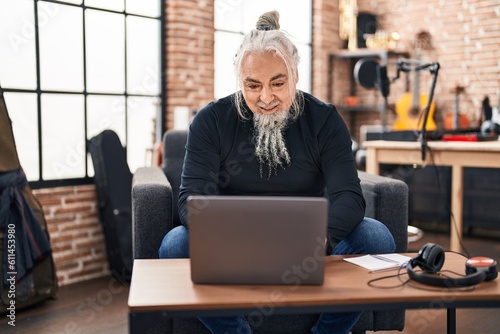 Middle age grey-haired man musician using laptop sitting on chair at music studio