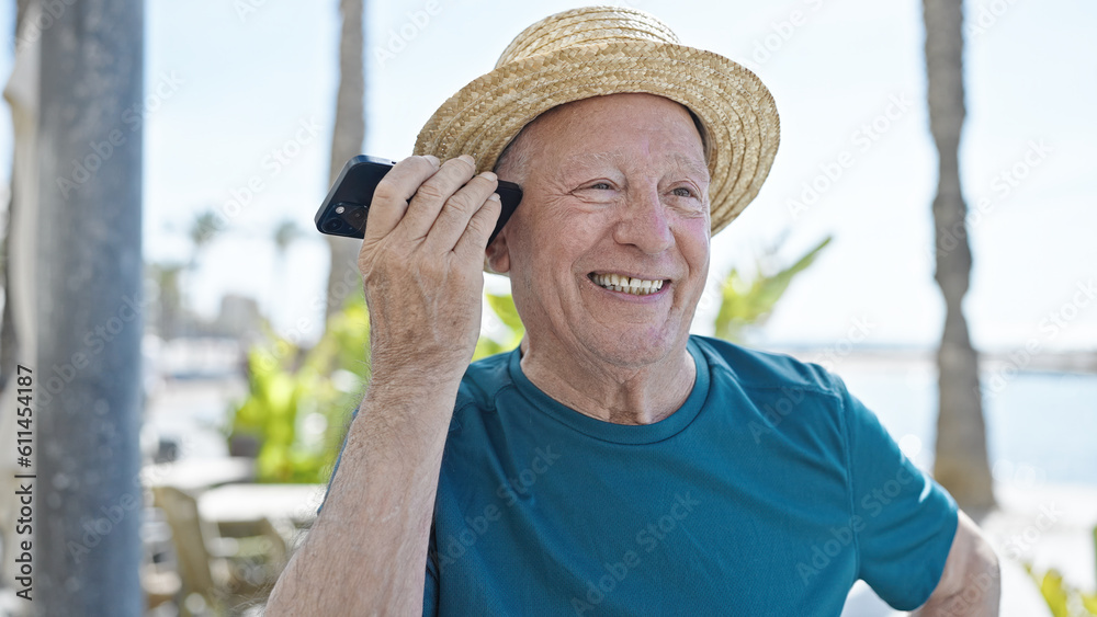 Senior grey-haired man tourist wearing summer hat listening voice message by smartphone at seaside