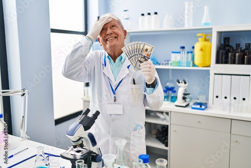 Senior scientist with grey hair working at laboratory holding dollars stressed and frustrated with hand on head  surprised and angry face