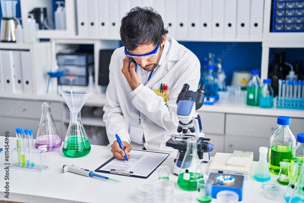 Fototapeta premium Young hispanic man scientist talking on the smartphone write on document at laboratory