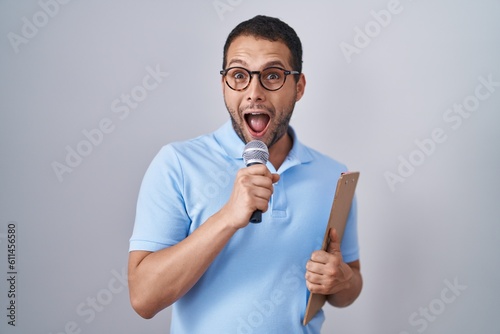 Hispanic man holding reporter microphone and clipboard afraid and shocked with surprise and amazed expression, fear and excited face.