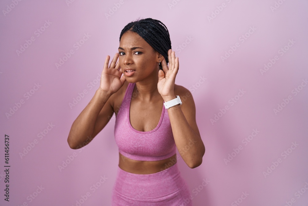 African american woman with braids wearing sportswear over pink background trying to hear both hands on ear gesture, curious for gossip. hearing problem, deaf African american woman with braids wearing sportswear over pink background trying to hear both hands on ear gesture, curious for gossip. hearing problem, deaf