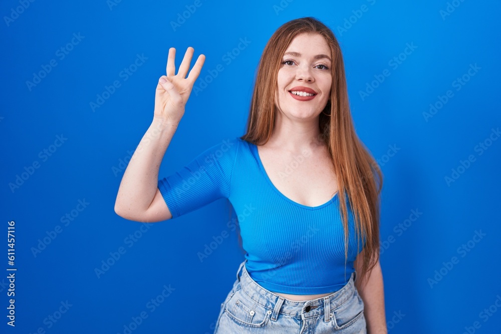 Redhead woman standing over blue background showing and pointing up with fingers number three while smiling confident and happy. Redhead woman standing over blue background showing and pointing up with fingers number three while smiling confident and happy.