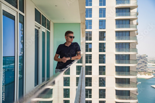 Attractive young man in sunglasses on skyscraper balcony with view of Dubai UAE, pensive looking away. Confident guy pondering on terrace of tower block. Recreation and leisure activity. Copy ad space