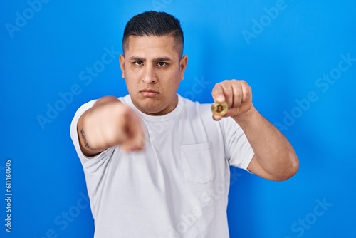 Hispanic young man holding virtual currency bitcoin pointing with finger to the camera and to you, confident gesture looking serious © Krakenimages.com
