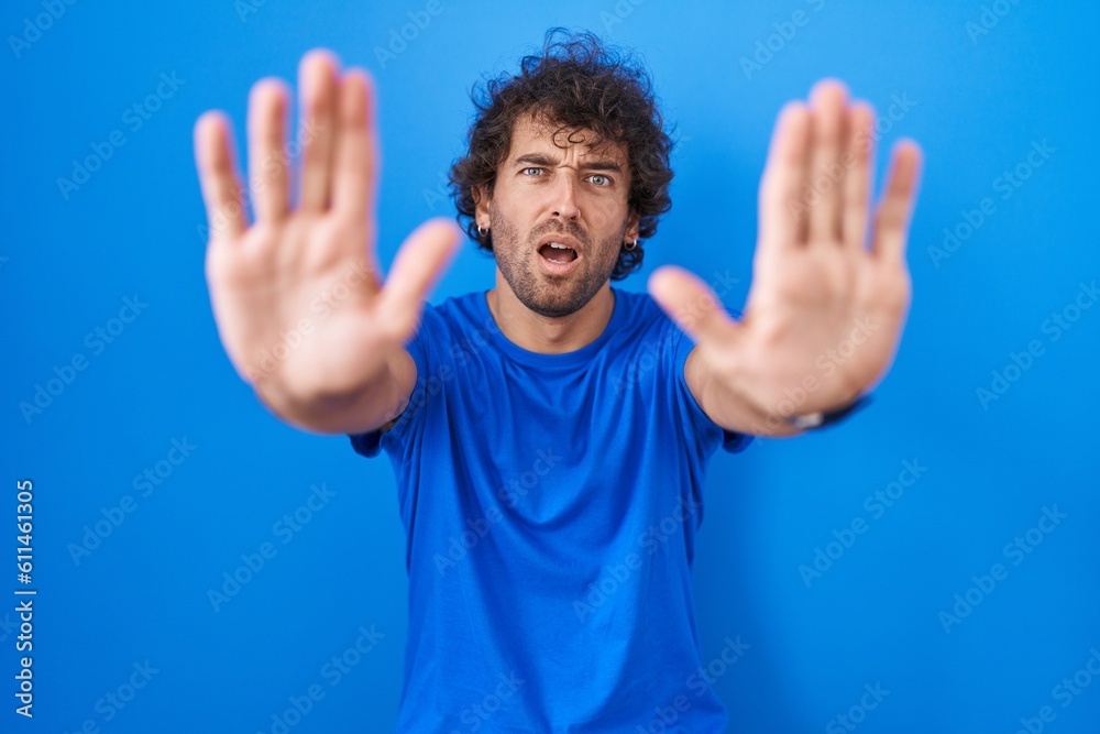 Hispanic young man standing over blue background doing stop gesture with hands palms, angry and frustration expression Hispanic young man standing over blue background doing stop gesture with hands palms, angry and frustration expression