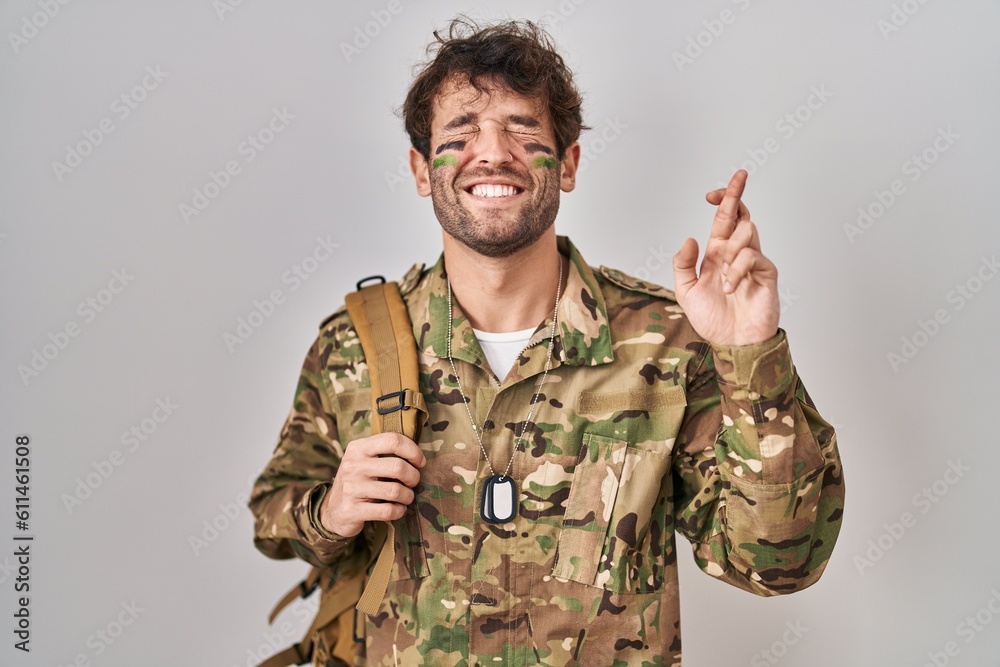 Hispanic young man wearing camouflage army uniform gesturing finger crossed smiling with hope and eyes closed. luck and superstitious concept.