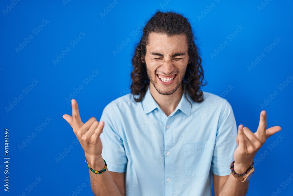 Young hispanic man standing over blue background shouting with crazy expression doing rock symbol with hands up. music star. heavy music concept. Young hispanic man standing over blue background shouting with crazy expression doing rock symbol with hands up. music star. heavy music concept.