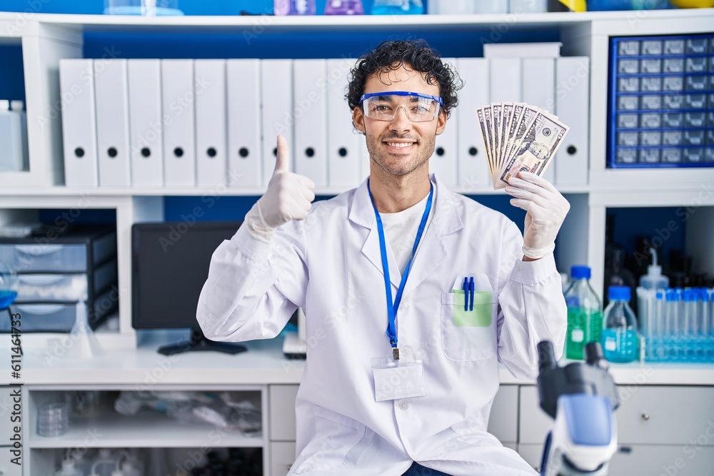 Hispanic man working at scientist laboratory holding money smiling happy and positive, thumb up doing excellent and approval sign Hispanic man working at scientist laboratory holding money smiling happy and positive, thumb up doing excellent and approval sign