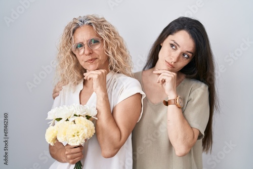 Mother and daughter holding bouquet of white flowers with hand on chin thinking about question, pensive expression. smiling with thoughtful face. doubt concept.