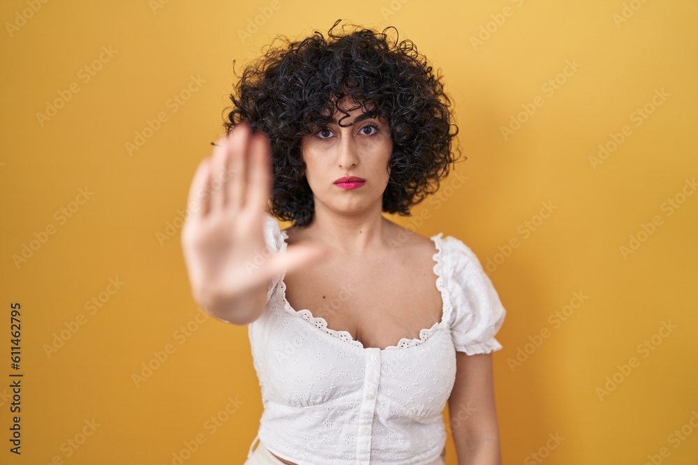 Young brunette woman with curly hair standing over yellow background doing stop sing with palm of the hand. warning expression with negative and serious gesture on the face. Young brunette woman with curly hair standing over yellow background doing stop sing with palm of the hand. warning expression with negative and serious gesture on the face.