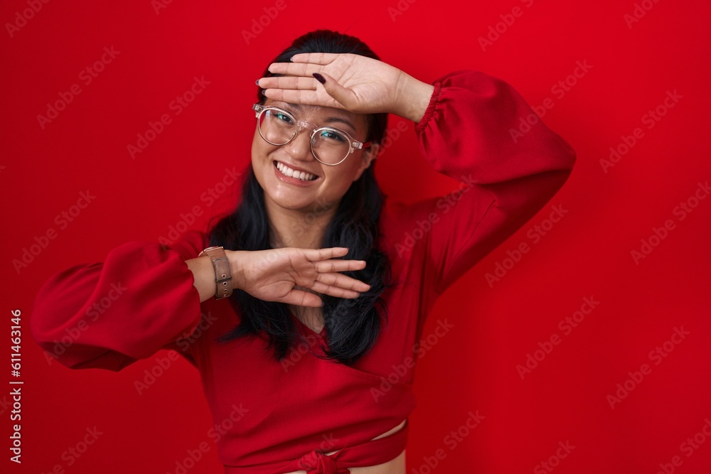 Asian young woman standing over red background smiling cheerful playing peek a boo with hands showing face. surprised and exited