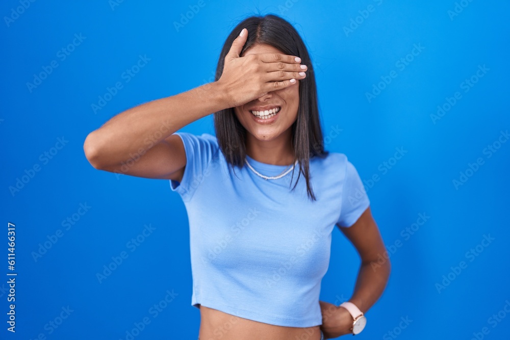 Brunette young woman standing over blue background smiling and laughing with hand on face covering eyes for surprise. blind concept. Brunette young woman standing over blue background smiling and laughing with hand on face covering eyes for surprise. blind concept.