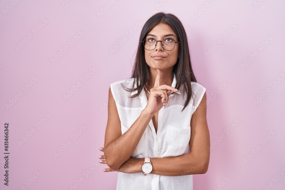 Brunette young woman standing over pink background wearing glasses thinking concentrated about doubt with finger on chin and looking up wondering Brunette young woman standing over pink background wearing glasses thinking concentrated about doubt with finger on chin and looking up wondering