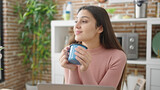 Young beautiful hispanic woman using laptop smelling cup of coffee at dinning room