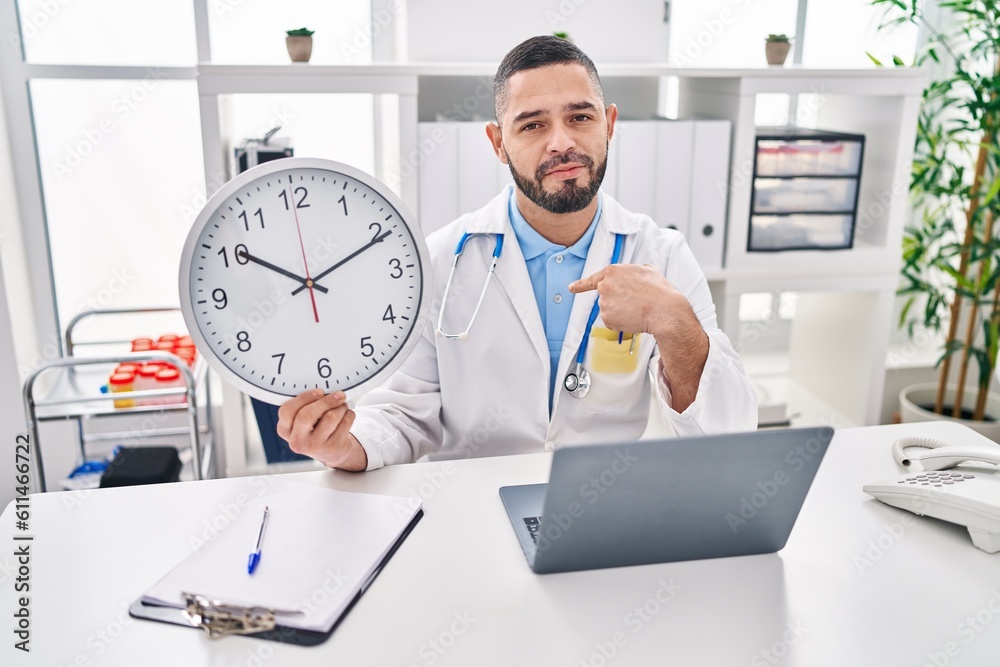 Hispanic doctor man holding clock at the clinic pointing finger to one self smiling happy and proud