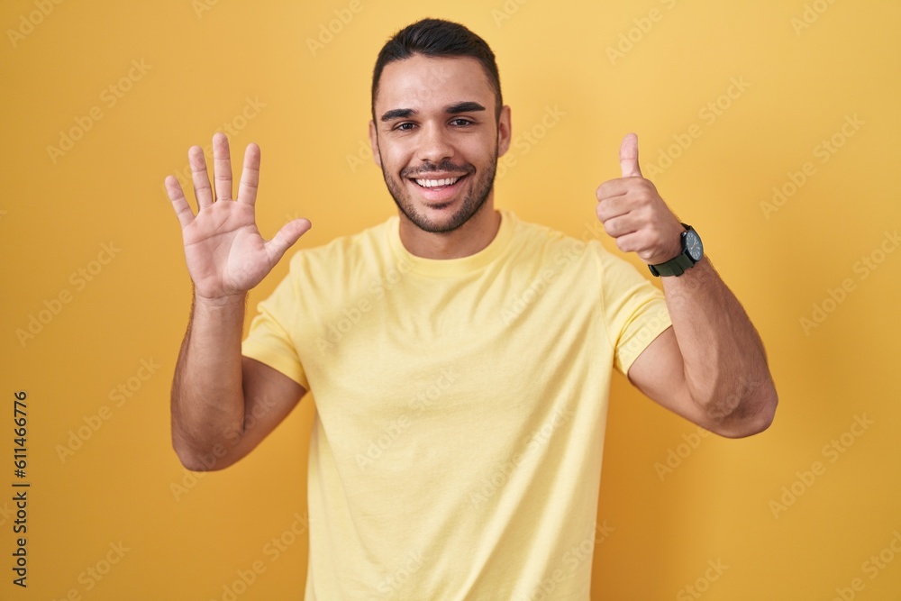 Young hispanic man standing over yellow background showing and pointing up with fingers number six while smiling confident and happy. Young hispanic man standing over yellow background showing and pointing up with fingers number six while smiling confident and happy.