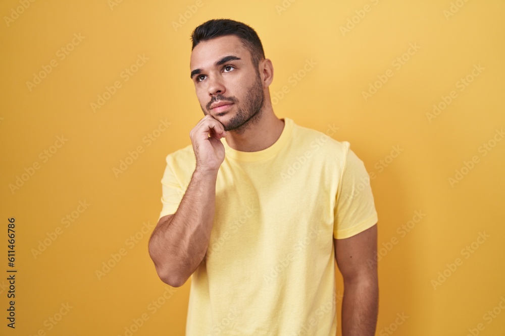 Young hispanic man standing over yellow background with hand on chin thinking about question, pensive expression. smiling with thoughtful face. doubt concept. Young hispanic man standing over yellow background with hand on chin thinking about question, pensive expression. smiling with thoughtful face. doubt concept.