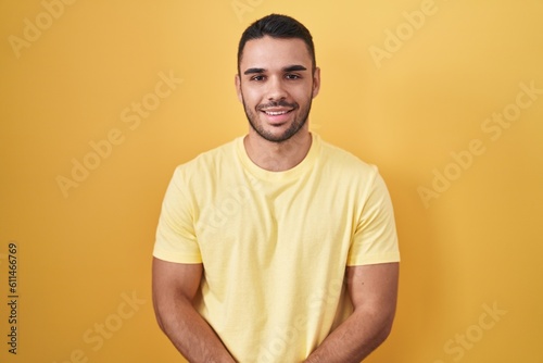 Young hispanic man standing over yellow background with hands together and crossed fingers smiling relaxed and cheerful. success and optimistic