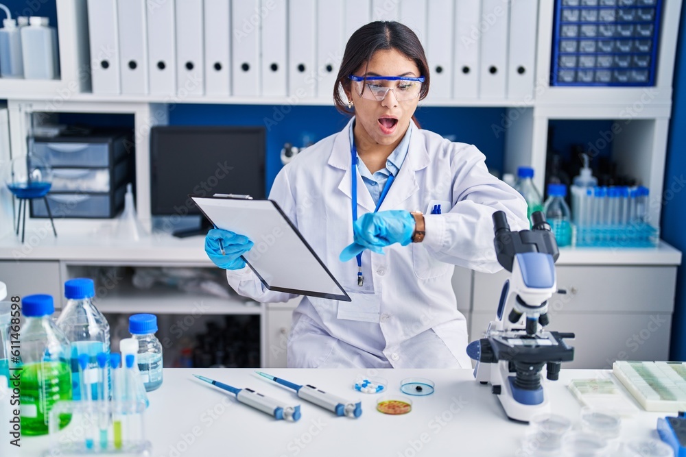 Hispanic young woman working at scientist laboratory looking at the watch time worried, afraid of getting late