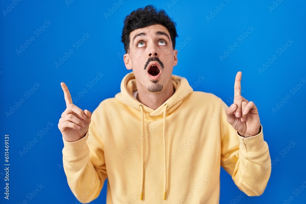 Hispanic man standing over blue background amazed and surprised looking up and pointing with fingers and raised arms. Hispanic man standing over blue background amazed and surprised looking up and pointing with fingers and raised arms.
