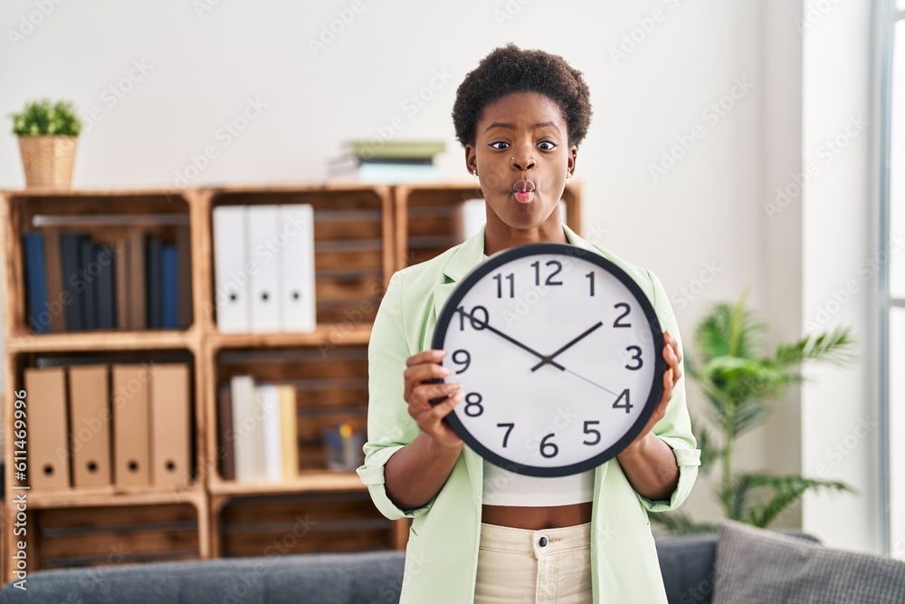 African american woman holding big clock making fish face with mouth and squinting eyes, crazy and comical. African american woman holding big clock making fish face with mouth and squinting eyes, crazy and comical.