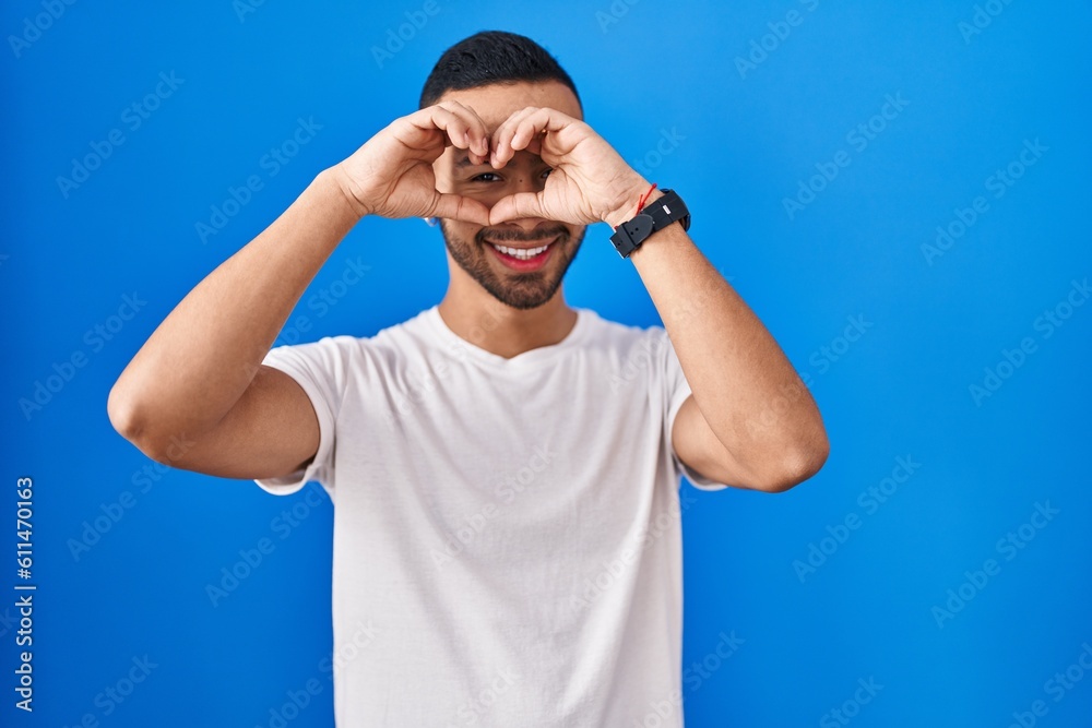 Naklejka premium Young hispanic man standing over blue background doing heart shape with hand and fingers smiling looking through sign
