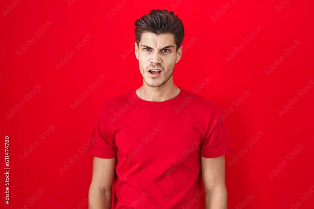 Young hispanic man standing over red background in shock face, looking skeptical and sarcastic, surprised with open mouth Young hispanic man standing over red background in shock face, looking skeptical and sarcastic, surprised with open mouth