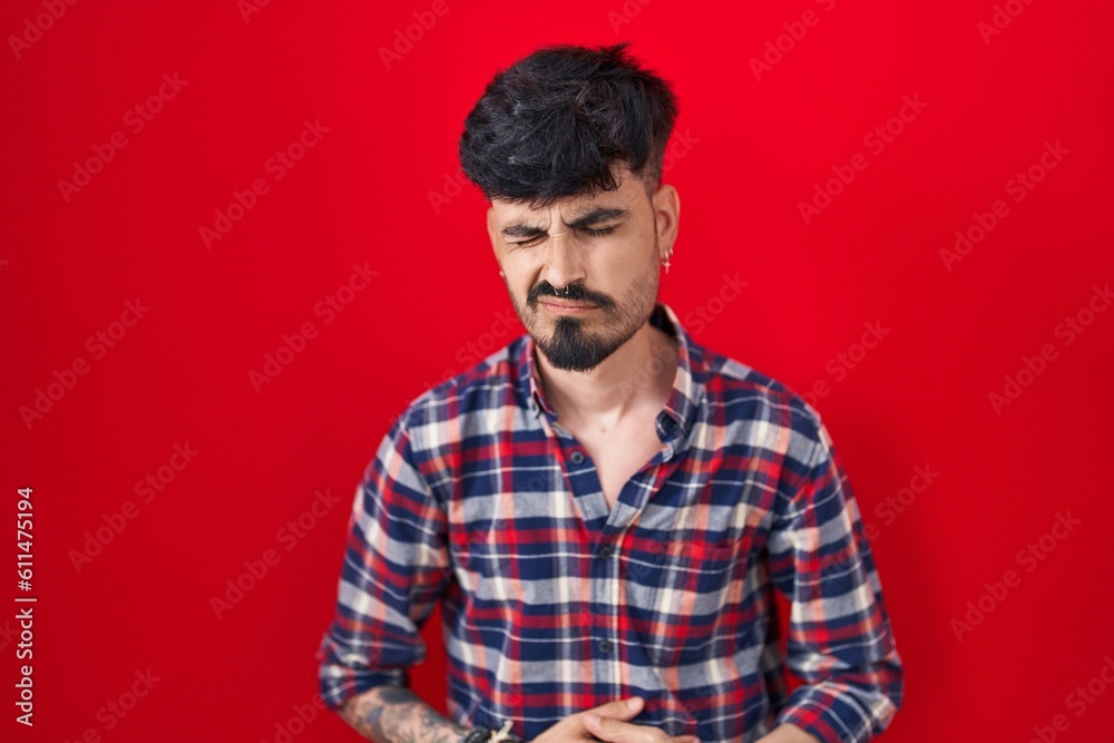 Young hispanic man with beard standing over red background with hand on stomach because indigestion, painful illness feeling unwell. ache concept. Young hispanic man with beard standing over red background with hand on stomach because indigestion, painful illness feeling unwell. ache concept.