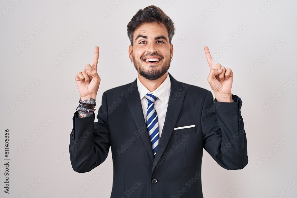Young hispanic man with tattoos wearing business suit and tie smiling amazed and surprised and pointing up with fingers and raised arms. Young hispanic man with tattoos wearing business suit and tie smiling amazed and surprised and pointing up with fingers and raised arms.