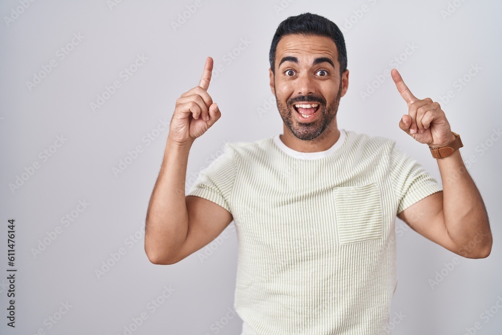 Hispanic man with beard standing over isolated background smiling amazed and surprised and pointing up with fingers and raised arms. Hispanic man with beard standing over isolated background smiling amazed and surprised and pointing up with fingers and raised arms.