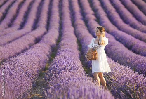 Fototapeta Naklejka Na Ścianę i Meble -  Full body portrait of young brunette woman from in casual white cotton dress enjoying beautiful lavender field and holding woven rattan fashion bag at sunny day