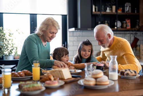 Small lovely boy and girl having rich delicious breakfast in the morning with grandmother and grandfather in the kitchen at home.