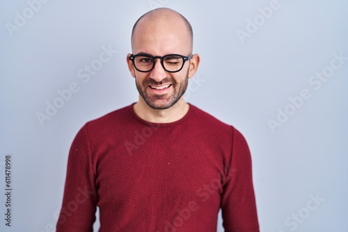 Young bald man with beard standing over white background wearing glasses winking looking at the camera with sexy expression, cheerful and happy face. © Krakenimages.com