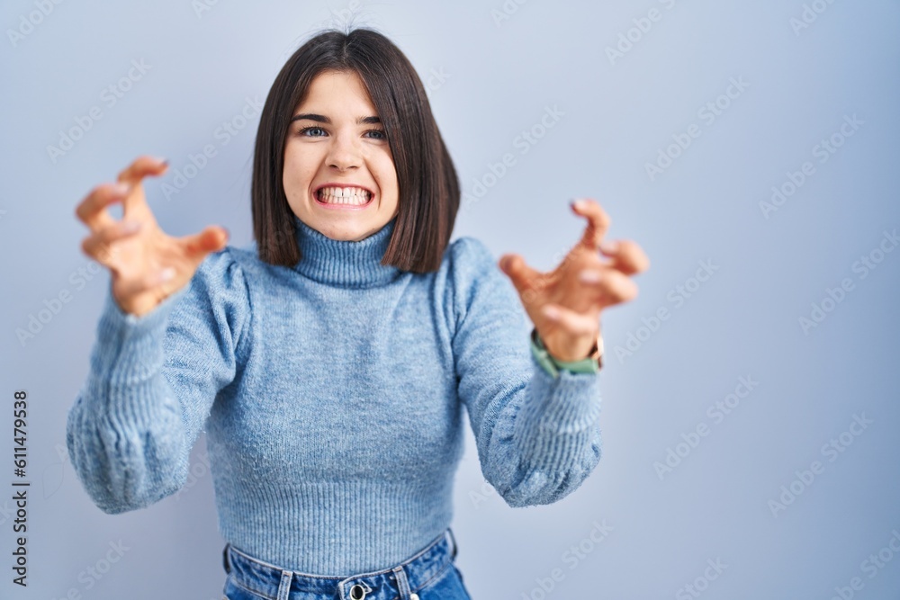 Young hispanic woman standing over blue background shouting frustrated with rage, hands trying to strangle, yelling mad Young hispanic woman standing over blue background shouting frustrated with rage, hands trying to strangle, yelling mad