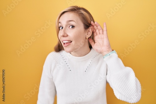 Young caucasian woman wearing white sweater over yellow background smiling with hand over ear listening an hearing to rumor or gossip. deafness concept.