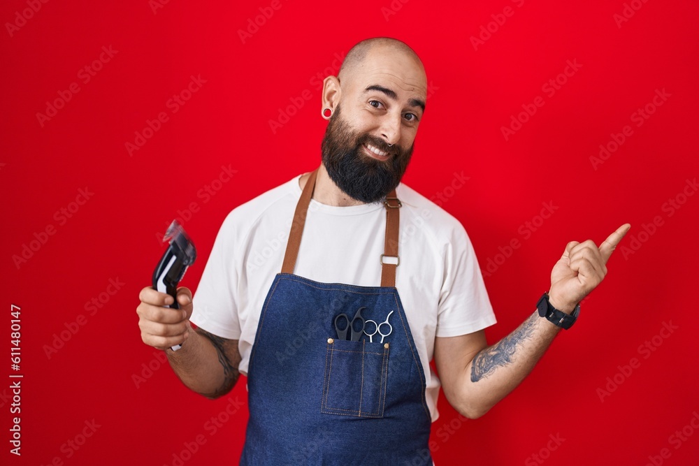 Young hispanic man with beard and tattoos wearing barber apron holding razor with a big smile on face, pointing with hand finger to the side looking at the camera. Young hispanic man with beard and tattoos wearing barber apron holding razor with a big smile on face, pointing with hand finger to the side looking at the camera.