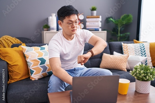 Young asian man using laptop at home sitting on the sofa looking at the watch time worried, afraid of getting late © Krakenimages.com